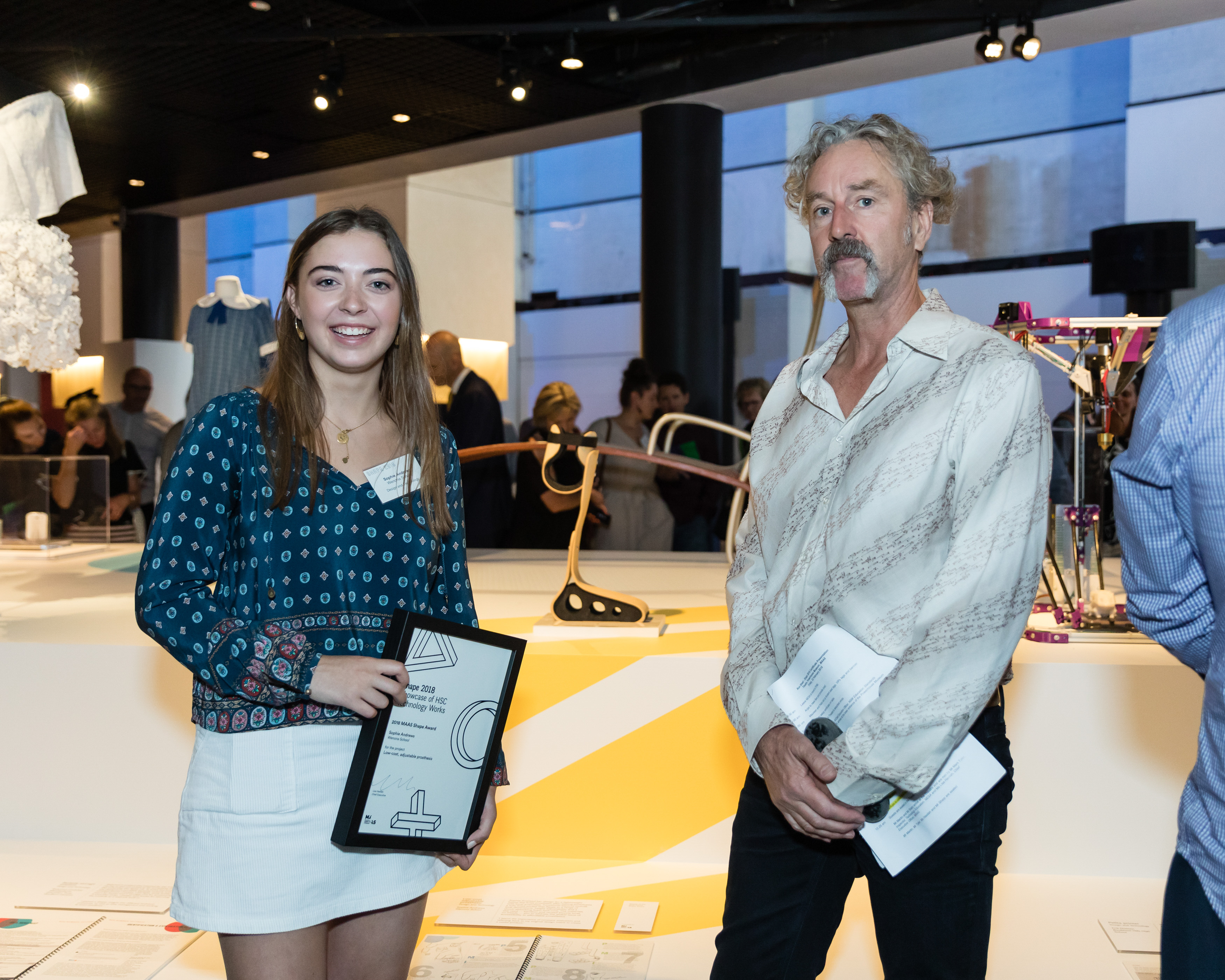 A young woman holding a certificate and a grey-haired man pose in front of an exhibition space.